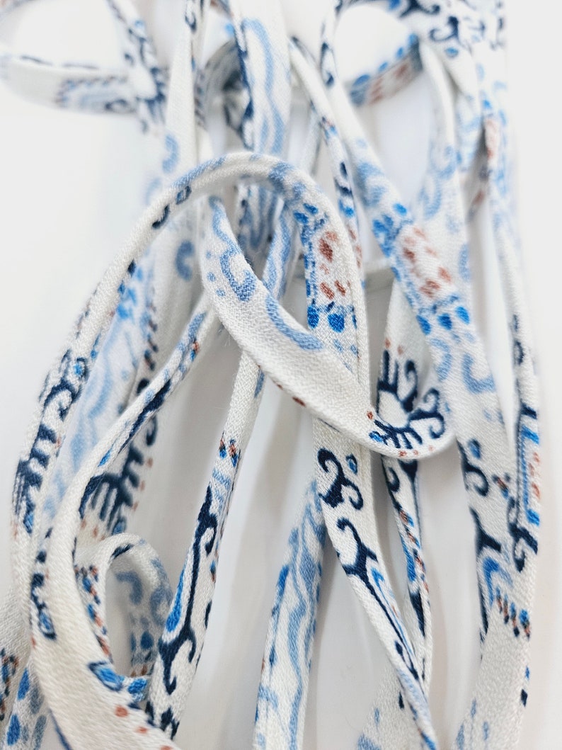 May include: A long, thin fabric ribbon or shoelace featuring a white base color with a repeating pattern of abstract shapes and lines in shades of dark blue, light blue, and brown. The material has a woven texture and is coiled in a loose pile.