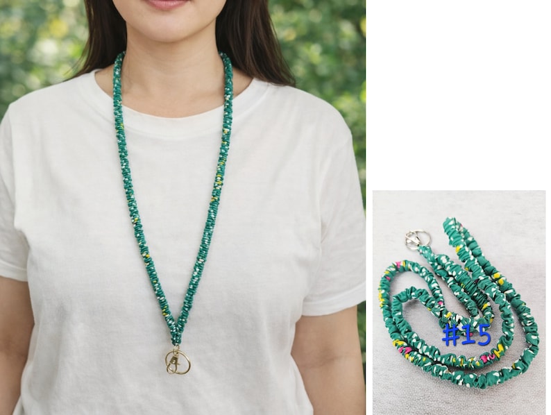 K&ouml;nnte beinhalten: Ein Stoff-Lanyard mit einer blaugr&uuml;nen Grundfarbe und einem mehrfarbigen Blumenmuster mit wei&szlig;en, gelben und rosa Akzenten. Das Lanyard verf&uuml;gt &uuml;ber einen goldfarbenen Karabinerhaken und einen Schl&uuml;sselring. Die Nummer 15 ist blau aufgedruckt.