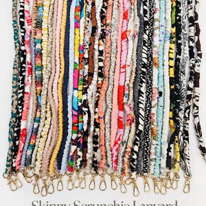 May include: A collection of skinny scrunchie lanyards in various colors and patterns, including floral, animal print, and solid hues. Each fabric-covered elastic strap features a gold-tone metal lobster clasp at the bottom end for attachment.