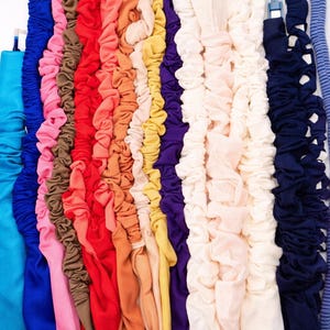 May include: A collection of colorful scrunchies in various shades, including blue, pink, red, orange, yellow, purple, and white. The scrunchies are made of fabric and are arranged vertically against a white background. The image showcases a variety of hair accessories.