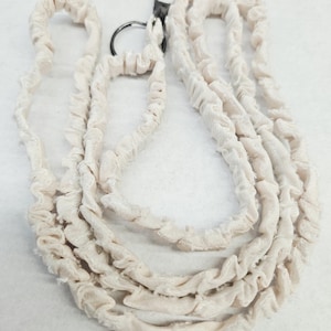 K&ouml;nnte beinhalten: Ein cremefarbenes Stoff-Lanyard mit einer gerafften, ger&uuml;schten Textur entlang der gesamten L&auml;nge des Bandes. Das Lanyard verf&uuml;gt &uuml;ber einen dunklen, runden Metallring und einen passenden Karabinerverschluss aus Metall zur Befestigung.