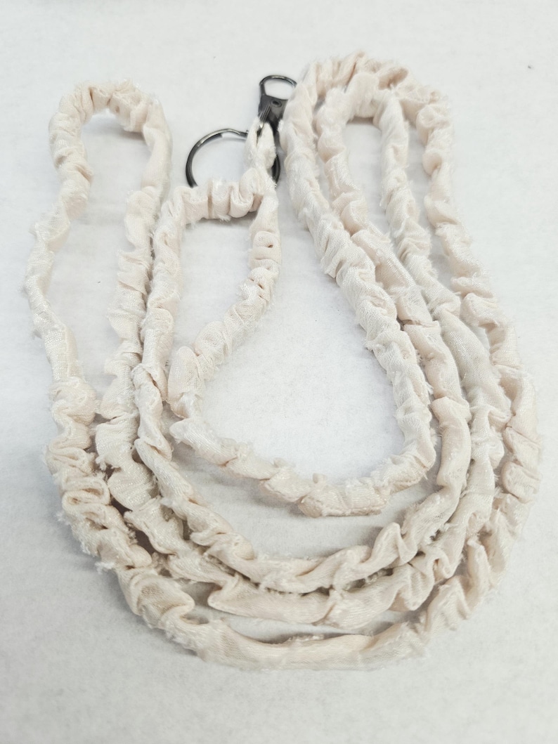 K&ouml;nnte beinhalten: Ein cremefarbenes Stoff-Lanyard mit einer gerafften, ger&uuml;schten Textur entlang der gesamten L&auml;nge des Bandes. Das Lanyard verf&uuml;gt &uuml;ber einen dunklen, runden Metallring und einen passenden Karabinerverschluss aus Metall zur Befestigung.
