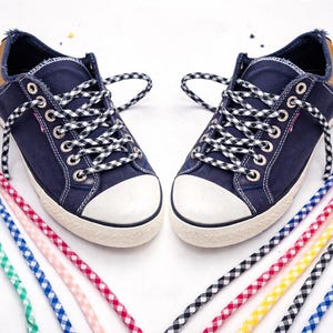 May include: A pair of navy blue canvas sneakers with white toe caps and brown leather accents. The shoes have black and white checkered laces. Colorful shoelaces in various patterns and colors are arranged around the shoes.