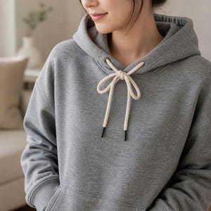 May include: A heather gray long-sleeve hooded sweatshirt featuring a large hood with thick, cream-colored braided drawstrings tied in a bow at the center. The garment includes a front kangaroo pocket and ribbed cuffs on the long sleeves.