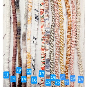 Op de afbeelding: Een collectie van veertien lange, dunne, met stof beklede elastische koorden die verticaal zijn gerangschikt. De koorden hebben verschillende kleuren en patronen, waaronder effen wit, bruin, grijs, geel, bloemenprints en geometrische ontwerpen, elk voorzien van een blauw genummerd label.