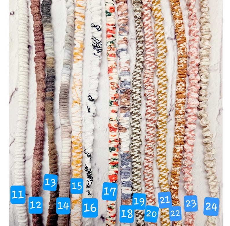 Op de afbeelding: Een collectie van veertien lange, dunne, met stof beklede elastische koorden die verticaal zijn gerangschikt. De koorden hebben verschillende kleuren en patronen, waaronder effen wit, bruin, grijs, geel, bloemenprints en geometrische ontwerpen, elk voorzien van een blauw genummerd label.