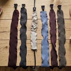 May include: An overhead shot displays a collection of fabric cord protectors in various colors, including burgundy, black, beige, blue, and gray. Each protector is attached to a black plug. A notebook, pen, and woven bag are also visible.