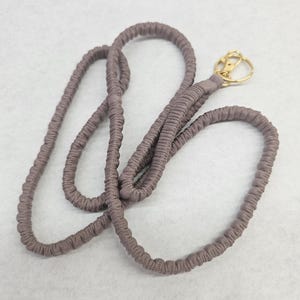 May include: A mauve-colored fabric lanyard featuring a gathered, elasticized texture throughout the cord. The lanyard includes a gold-toned metal swivel lobster clasp attached to one end, providing a functional connector for keys or identification badges.