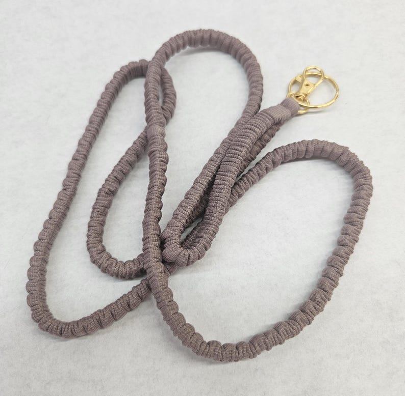 May include: A mauve-colored fabric lanyard featuring a gathered, elasticized texture throughout the cord. The lanyard includes a gold-toned metal swivel lobster clasp attached to one end, providing a functional connector for keys or identification badges.