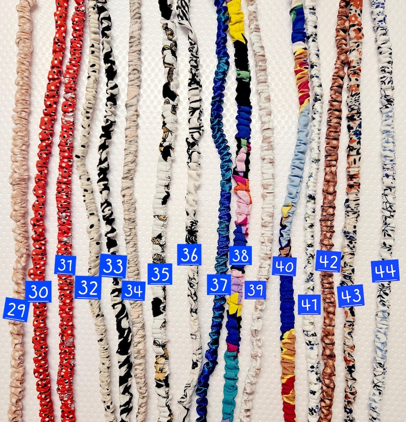 K&ouml;nnte beinhalten: Eine Sammlung von sechzehn stoffbezogenen elastischen Kordeln, die vertikal angeordnet sind. Jede Kordel weist unterschiedliche Muster und Farben auf, die mit blauen nummerierten Etiketten von 29 bis 44 versehen sind, darunter florale, abstrakte und geometrische Drucke.