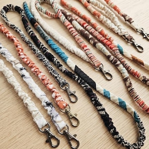 May include: A collection of fabric lanyards featuring various patterns and colors, including orange, black, white, and blue. Each lanyard is constructed from gathered fabric and is equipped with a metal swivel lobster clasp at the end.