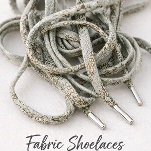 Puede incluir: Primer plano de cordones de tela con un estampado floral en tonos grises y marrones. Los cordones tienen punteras met&aacute;licas plateadas. El texto "Fabric Shoelaces" se muestra debajo.