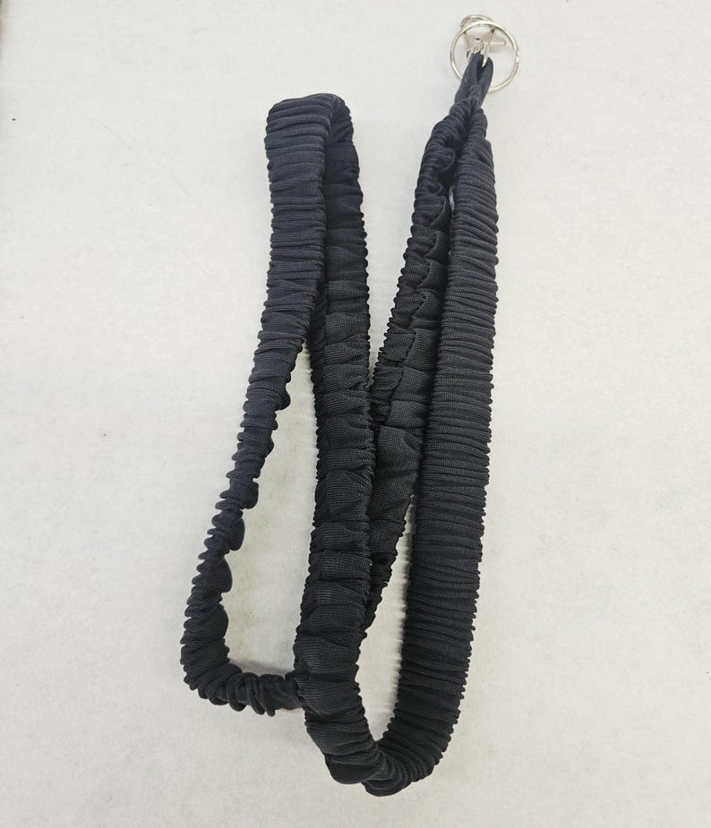 May include: A black fabric lanyard featuring a gathered, elasticized texture throughout the length of the strap. The lanyard is finished with a circular silver-toned metal split ring attachment at the top for securing keys or identification badges.