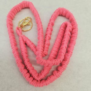 May include: A bright pink fabric lanyard featuring a gathered, scrunchie-style texture along the entire length of the cord. The lanyard is equipped with a circular gold-toned metal split ring and a matching gold-toned lobster claw clasp.