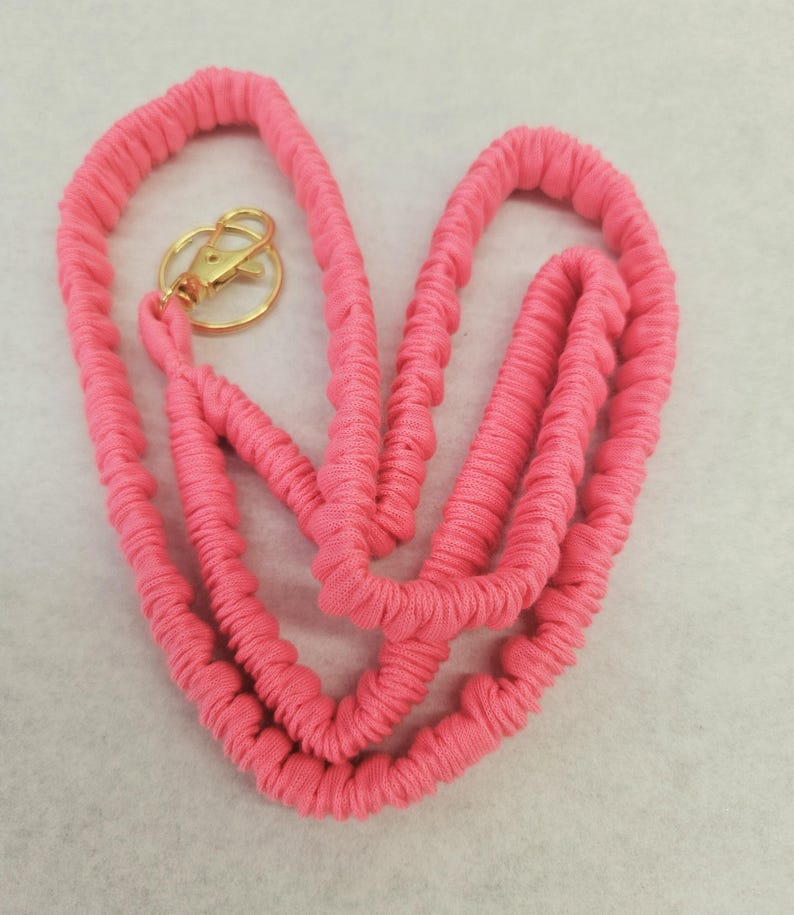 May include: A bright pink fabric lanyard featuring a gathered, scrunchie-style texture along the entire length of the cord. The lanyard is equipped with a circular gold-toned metal split ring and a matching gold-toned lobster claw clasp.