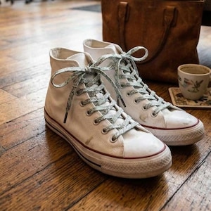 Puede incluir: Zapatillas altas blancas con cordones estampados y una franja roja en la suela. Los zapatos est&aacute;n sobre un suelo de madera, con un bolso de cuero y una taza de t&eacute; en el fondo.