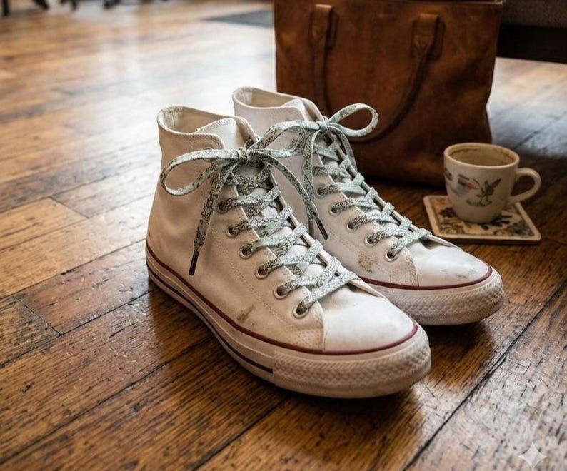 Puede incluir: Zapatillas altas blancas con cordones estampados y una franja roja en la suela. Los zapatos est&aacute;n sobre un suelo de madera, con un bolso de cuero y una taza de t&eacute; en el fondo.