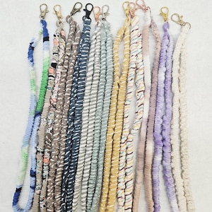 Op de afbeelding: Een collectie van twaalf stoffen koorden met diverse patronen en kleuren, waaronder blauw, groen, roze, geel, paars en neutrale tinten. Elk koord is aan de bovenzijde afgewerkt met een draaibare metalen karabijnsluiting.