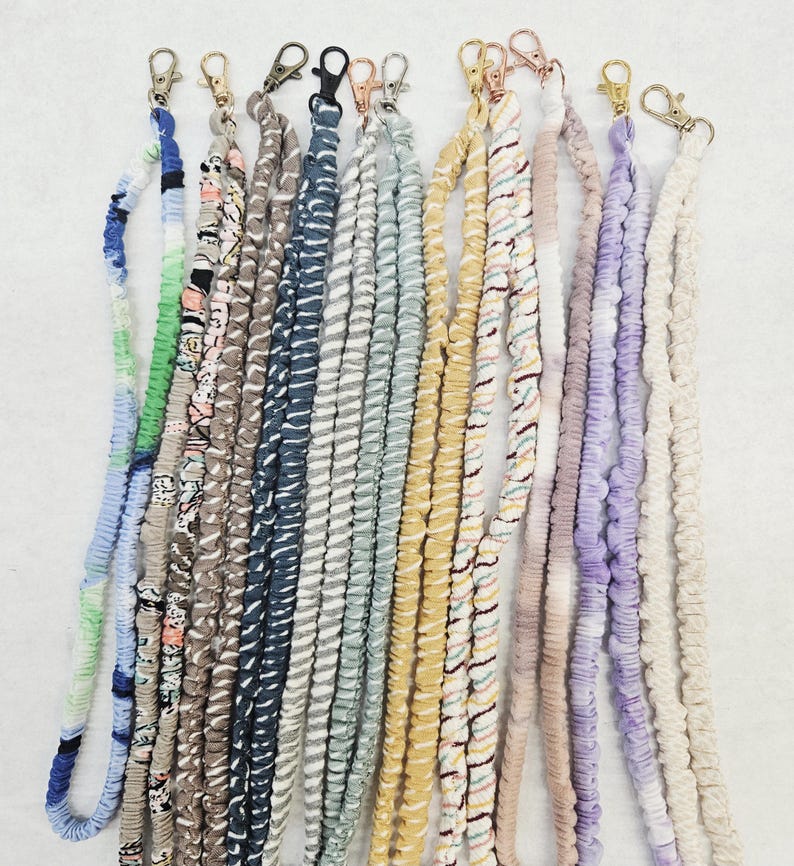 Op de afbeelding: Een collectie van twaalf stoffen koorden met diverse patronen en kleuren, waaronder blauw, groen, roze, geel, paars en neutrale tinten. Elk koord is aan de bovenzijde afgewerkt met een draaibare metalen karabijnsluiting.