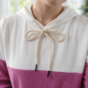 Puede incluir: Una sudadera con capucha de bloques de color en blanco y magenta. La mitad superior es blanca, la mitad inferior es magenta y las mangas son azules. La sudadera con capucha tiene un cordón con cuerda beige y punteras negras.