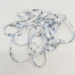May include: A pair of flat shoelaces featuring a white base with a repeating blue and light blue abstract pattern. The laces are constructed from a woven fabric material and are finished with silver-toned metal aglets at each of the ends.
