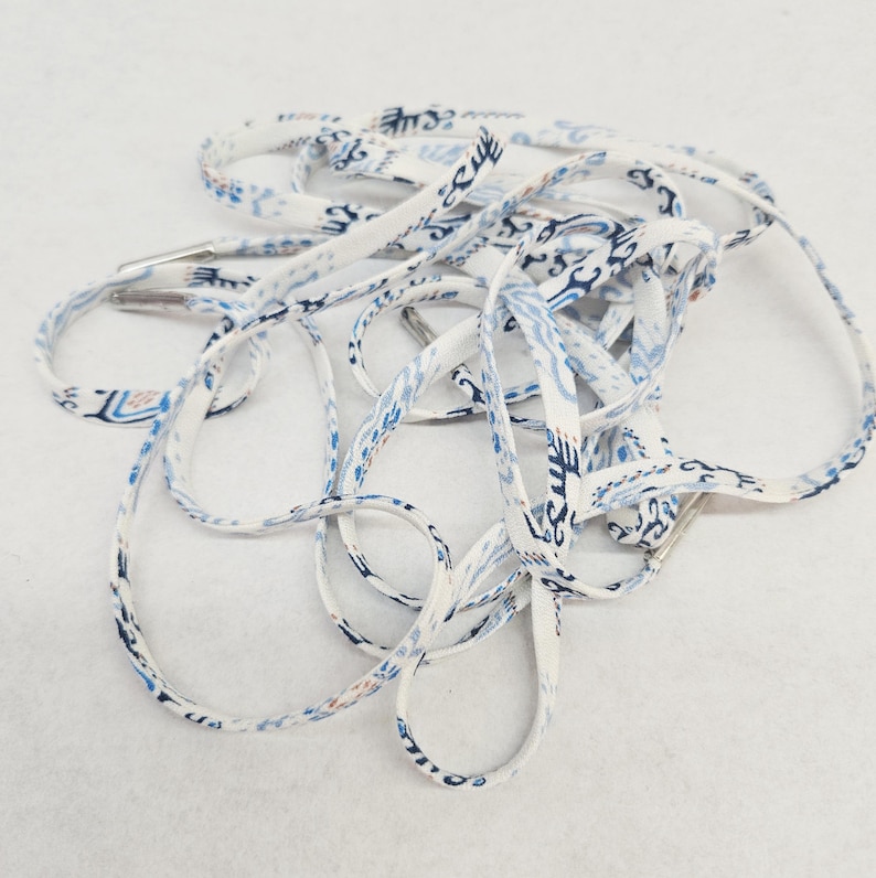 May include: A pair of flat shoelaces featuring a white base with a repeating blue and light blue abstract pattern. The laces are constructed from a woven fabric material and are finished with silver-toned metal aglets at each of the ends.