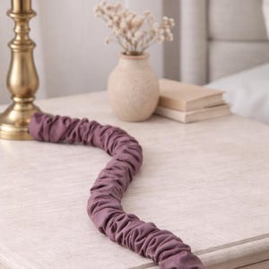 May include: A mauve fabric hair curler lies on a light-coloured bedside table. A gold-coloured lamp and a small vase with dried flowers are also on the table. A stack of books is visible in the background.