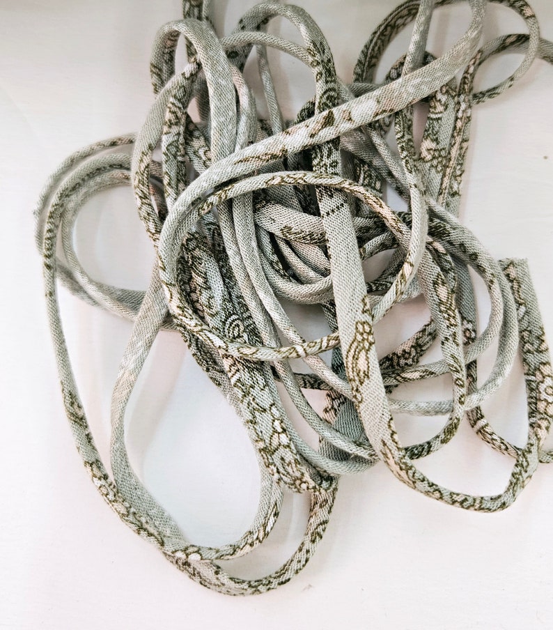 Puede incluir: Un haz enredado de cuerda gris claro con un patr&oacute;n floral repetido en verde oliva y blanco. La cuerda parece estar hecha de tela y est&aacute; enrollada, creando una apariencia texturizada.