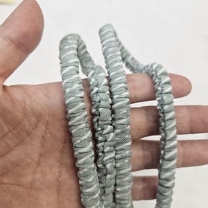 Op de afbeelding: Een haaraccessoire van saliegroen en wit gestreepte stof met een geplooid, elastisch ontwerp. Het materiaal heeft een zachte, getextureerde uitstraling met een herhalend diagonaal patroon van dunne witte lijnen op een gedempte groene achtergrond.