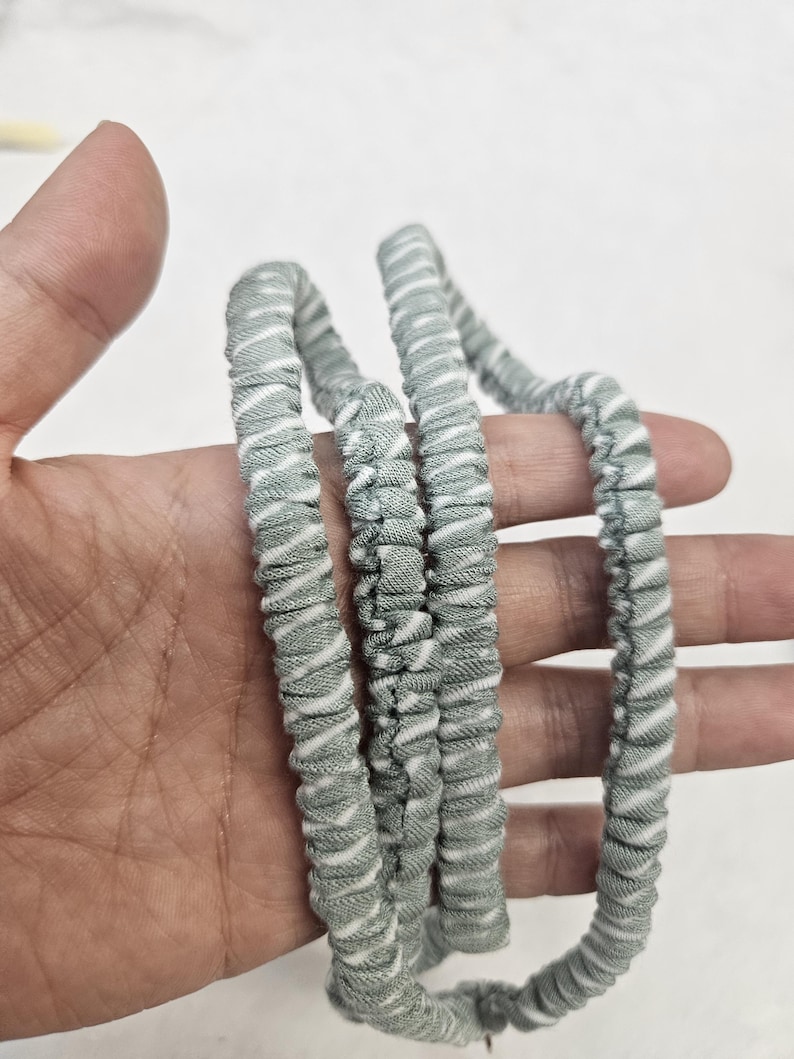Op de afbeelding: Een haaraccessoire van saliegroen en wit gestreepte stof met een geplooid, elastisch ontwerp. Het materiaal heeft een zachte, getextureerde uitstraling met een herhalend diagonaal patroon van dunne witte lijnen op een gedempte groene achtergrond.