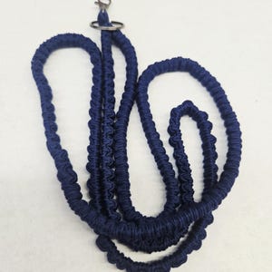 May include: A dark blue lanyard featuring a braided cord design with a textured, knotted appearance. The lanyard includes a silver-toned metal lobster clasp and a circular split ring at the top for attaching items, presented in a coiled arrangement.
