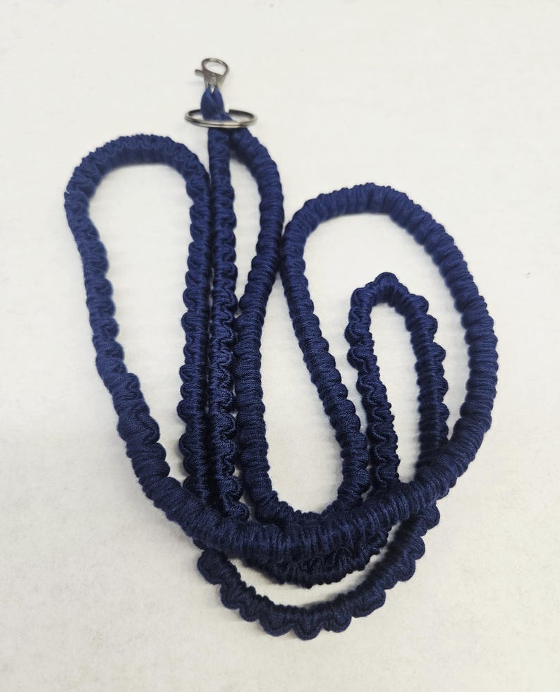 May include: A dark blue lanyard featuring a braided cord design with a textured, knotted appearance. The lanyard includes a silver-toned metal lobster clasp and a circular split ring at the top for attaching items, presented in a coiled arrangement.