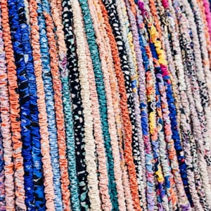 May include: Close-up of a colorful textile with vertical stripes. The stripes feature various patterns and colors, including blue, orange, white, and black. The textile appears to be made of fabric strips, possibly for a rug or decorative item.