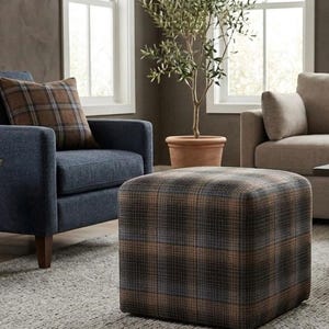 May include: A living room with a blue armchair and a beige sofa. A plaid ottoman is in front of the armchair. A brown plaid cushion sits on the armchair. A small wooden table and a potted olive tree are also present.