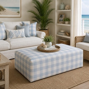 Può includere: Un pouf a quadretti blu chiaro e bianco è posto di fronte a un divano bianco con cuscini abbinati. Un vassoio intrecciato con oggetti decorativi è posto sopra. La stanza ha un tema costiero.