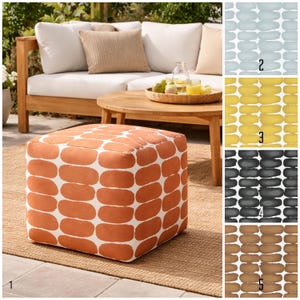 Könnte beinhalten: Ein quadratischer Hocker mit einem weißen und orangefarbenen ovalen Muster. Der Hocker steht auf einem gewebten Teppich, mit einem Holzsofa und einem Couchtisch im Hintergrund. Das Sofa hat weiße Kissen und ein beiges Kissen.