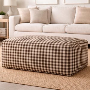 Puede incluir: Un otomán rectangular con un patrón de cuadros marrones y beige se encuentra sobre una alfombra de yute tejida. El otomán está frente a un sofá color crema con cojines beige. También se ven una mesa auxiliar y una cesta tejida.