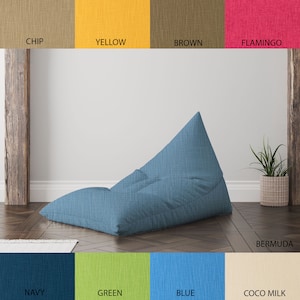 May include: A blue bean bag chair in a wedge shape. The chair is on a wooden floor with a light blue rug in the foreground. The background is a white wall with a wooden beam on the left side of the image. The image also includes a small potted plant on the right side of the image.