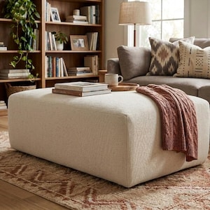 Può includere: Un grande pouf rettangolare di colore crema neutro, con un tessuto strutturato. Una coperta di lana a maglia di colore ruggine è appoggiata sul pouf. Un vassoio di legno con una tazza è sopra. Il pouf è in un salotto.