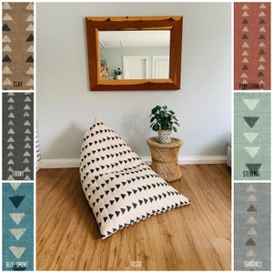 May include: A white and black patterned bean bag chair with a triangular design. The chair is sitting on a wooden floor in a room with a mirror and a plant. The chair is labeled "Fossil".