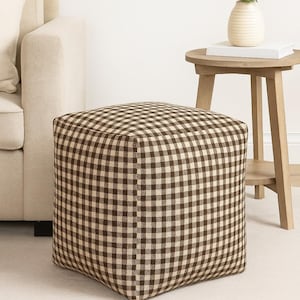 Può includere: Un pouf quadrato con un motivo a quadretti marrone e beige. Il poggiapiedi a forma di cubo è posto su un tappeto colorato chiaro. Un divano beige e un tavolino in legno con un vaso e un libro sono sullo sfondo.