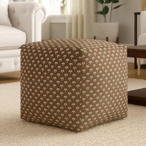 Può includere: Un pouf quadrato marrone con un motivo a fiocco bianco ripetuto. Il pouf a forma di cubo è posizionato su un tappeto beige chiaro. Un elemento d'arredo.
