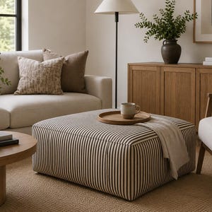 May include: A square ottoman upholstered in a dark brown and cream vertical striped fabric. The ottoman features a flat top surface holding a round wooden tray with a ceramic mug and a draped light-coloured textile, positioned in a living room.