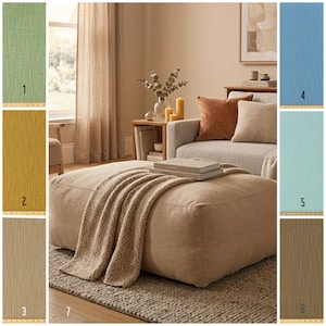 May include: A living room setting with a large beige ottoman, a folded beige blanket, and books. A grey sofa with brown and beige cushions, a wooden side table, and fabric swatches in green, yellow, blue, and brown are also visible. The room has a neutral colour palette.