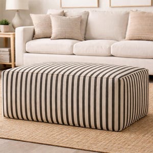 Peut inclure: Un pouf rectangulaire à rayures crème et noires. Le pouf est placé sur un tapis tissé devant un canapé blanc avec des coussins beiges. La pièce a une palette de couleurs neutres.