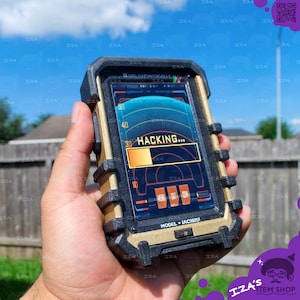 May include: A handheld electronic device with a rugged black and gold frame, featuring a digital screen that displays a radar-style interface with the text HACKING and a loading bar. The device is labeled with the text MODEL IAC2025I at the bottom.