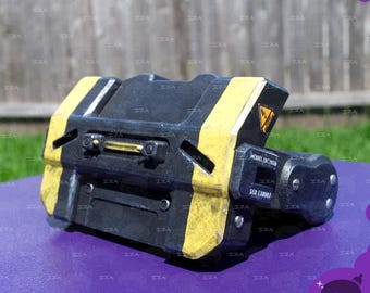 Pode incluir: Um acessório de estilo industrial preto e amarelo com acabamento desgastado, um símbolo de perigo triangular amarelo e texto impresso que diz MODEL IAC2026 e SER 130007. O objeto tem uma forma retangular com detalhes metálicos.