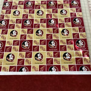 May include: A rectangular piece of fabric featuring a repeating pattern of the Florida State Seminoles logo in maroon, gold, and white. The fabric has a maroon border at the bottom and a white border at the top.