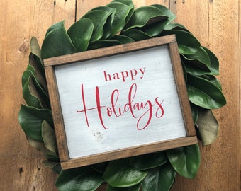 May include: A rectangular wooden sign with a white painted surface and a dark brown frame. The text "happy Holidays" is printed in red, with "happy" in a simple font and "Holidays" in a cursive script. The sign is centered on a green leaf wreath.