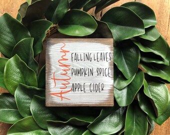 May include: A square wooden sign with the word "Autumn" in orange script, listing "Falling Leaves, Pumpkin Spice, Apple Cider" in black text. The sign is surrounded by a wreath of large, green leaves, set against a wooden background.